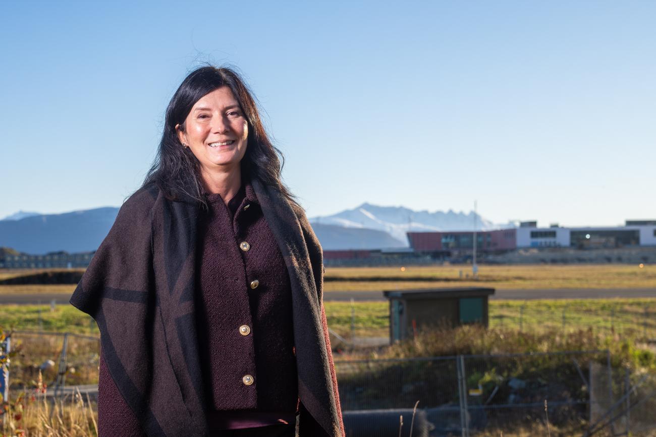 Monica Hansen Fjellstad i m&oslash;rk ullk&aring;pe st&aring;r utend&oslash;rs foran et gjerde og en gresslette, med bygninger og sn&oslash;dekte fjell i bakgrunnen